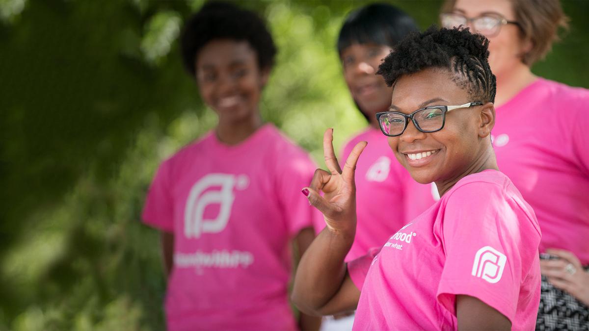 A Planned Parenthood volunteer smiles and holds up a peace sign while canvassing a neighborhood. She and others in the background all wear pink t-shirts.