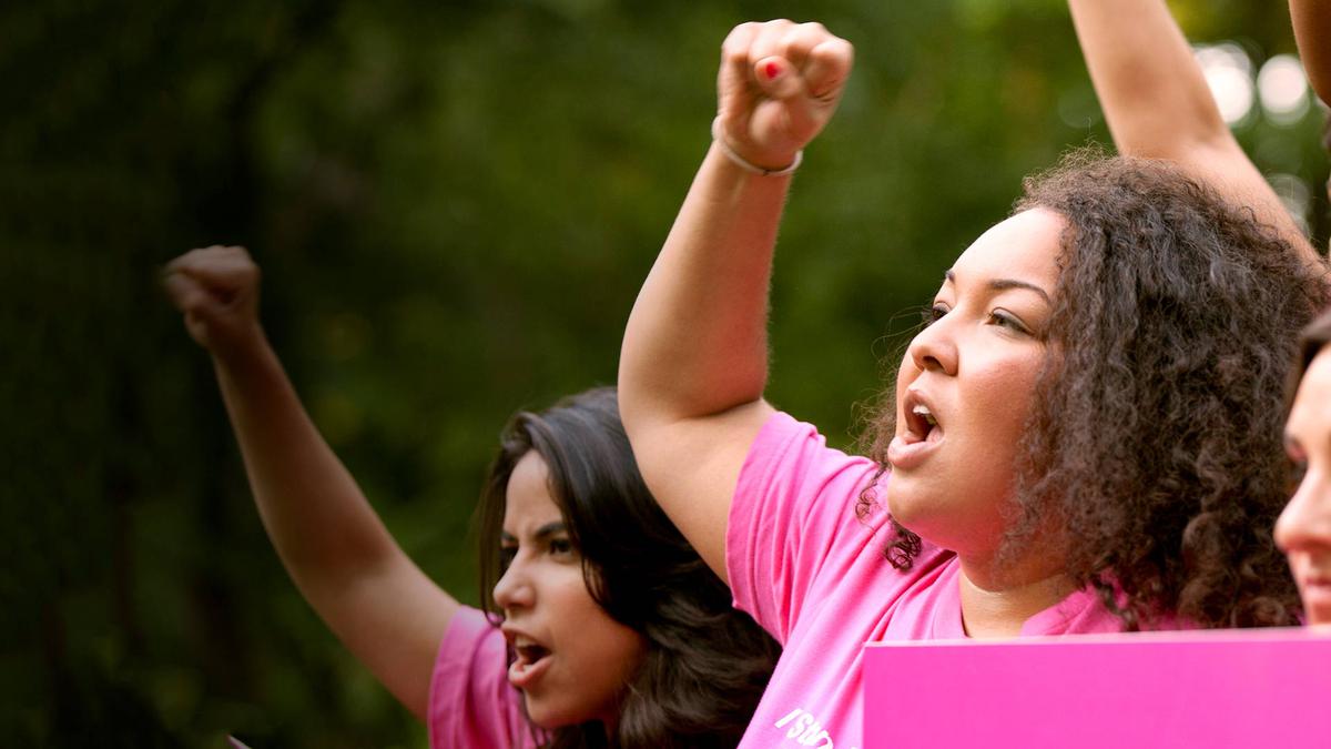 A person of color in their twenties chants at a rally for sexual and reproductive health wearing a pink t-shirt.