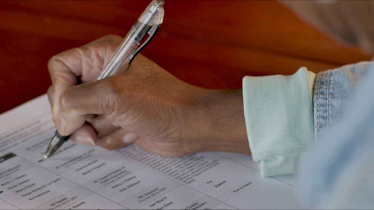 The hand of a person of color filling out their Minnesota ballot at their kitchen table.